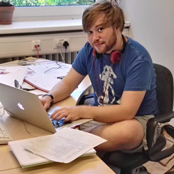 A photograph of me sitting at my desk during my time at DESY. My laptop is open and I am surrounded by papers.